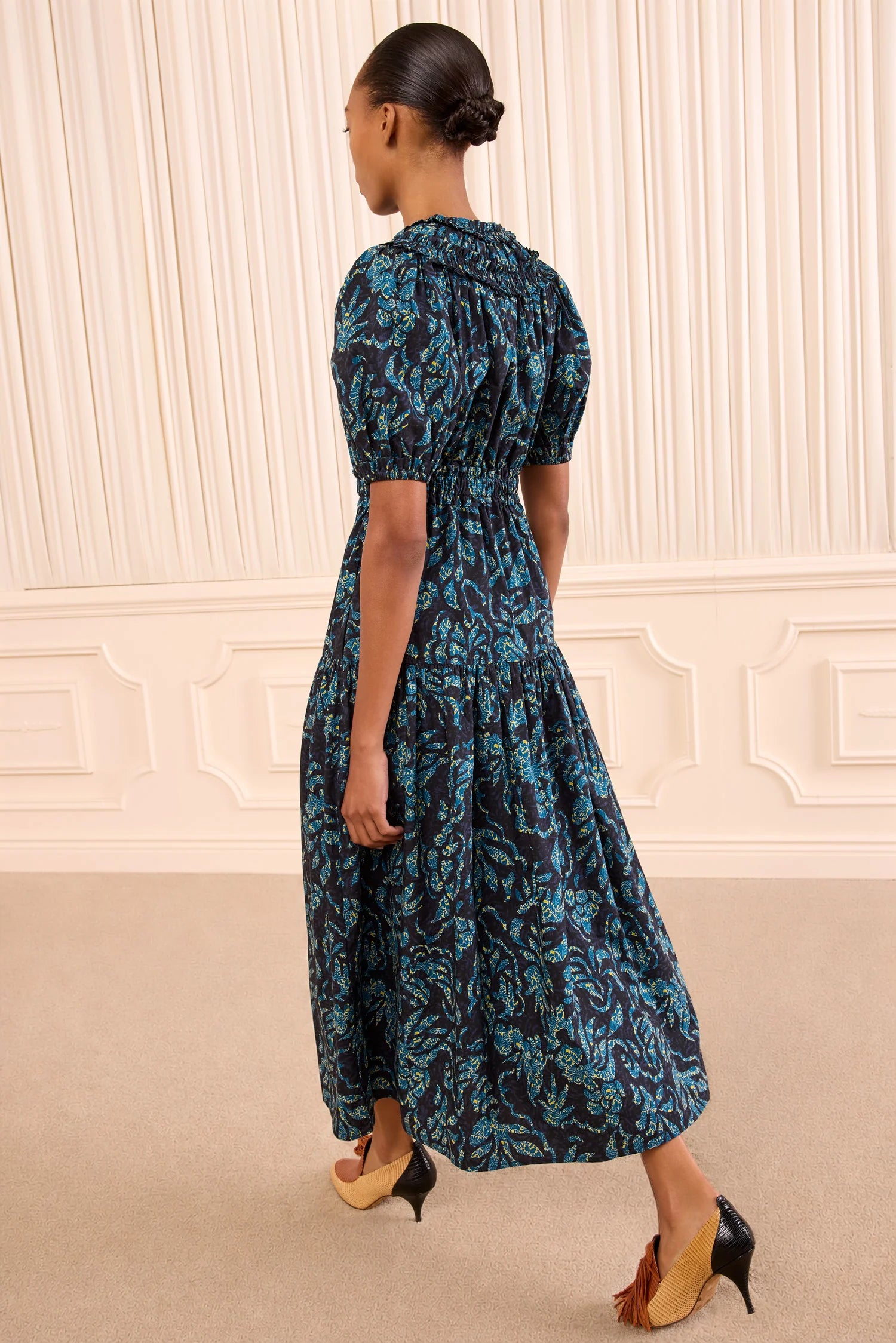 A woman with her hair in a bun wears the Othella Cotton Dress by Ulla Johnson—a dark blue floral print maxi dress with elegant puff sleeves—and black heeled shoes, standing in a beige room with decorative wall panels.