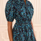 A woman stands indoors against a light-striped wall wearing the Ulla Johnson Othella Cotton Dress—a navy blue midi dress adorned with teal and yellow leaf patterns, puff sleeves, a gathered neckline, a shirred waist, and a pleated skirt.