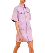 A person wearing a Wisteria lavender Ulla Johnson Painters Dress with a shirt-inspired silhouette, front pockets, and gray heels with bow details.