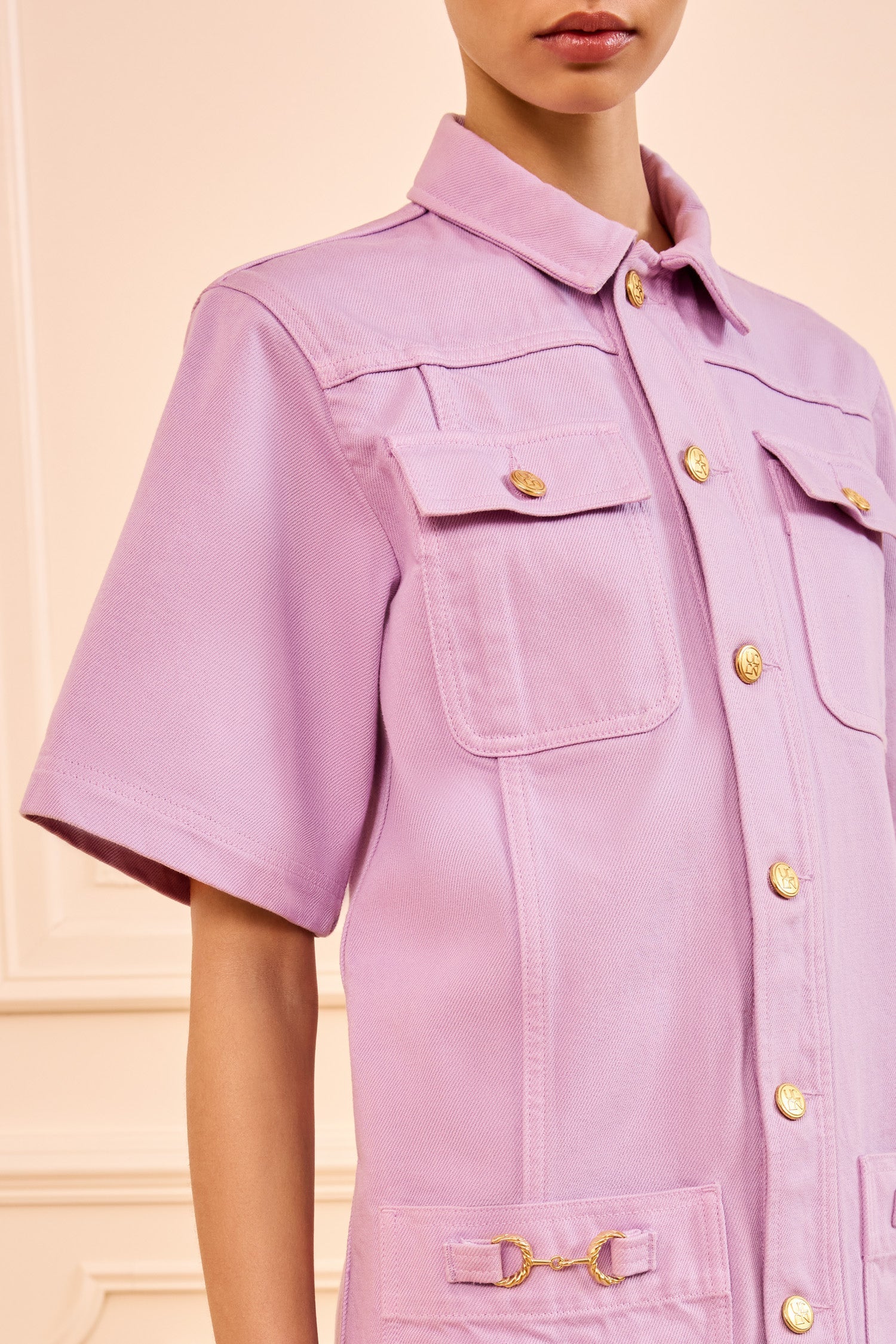 A person wears the Ulla Johnson Painters Dress—a short-sleeve, Wisteria lavender button-up style with gold buttons and front pockets—shown from shoulders to mid-thigh.
