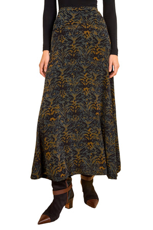 A person wearing the Ulla Johnson Paola Jersey Skirt, a long dark A-line style with a gold and blue floral design, paired with a black top and heeled boots. The person's head is not visible.