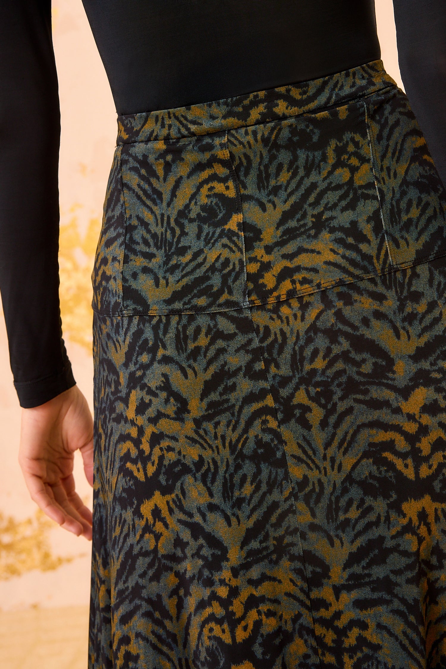 Someone stands facing away from the camera wearing a long-sleeve black tricot jersey top and the Ulla Johnson Paola Jersey Skirt, a high-waisted A-line skirt featuring a dark abstract leafy pattern.