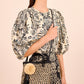 Woman in a floral blouse and skirt holds the Ulla Johnson Raffia Mini Crossbody, featuring a pom-pom and tassel, against a light striped background.
