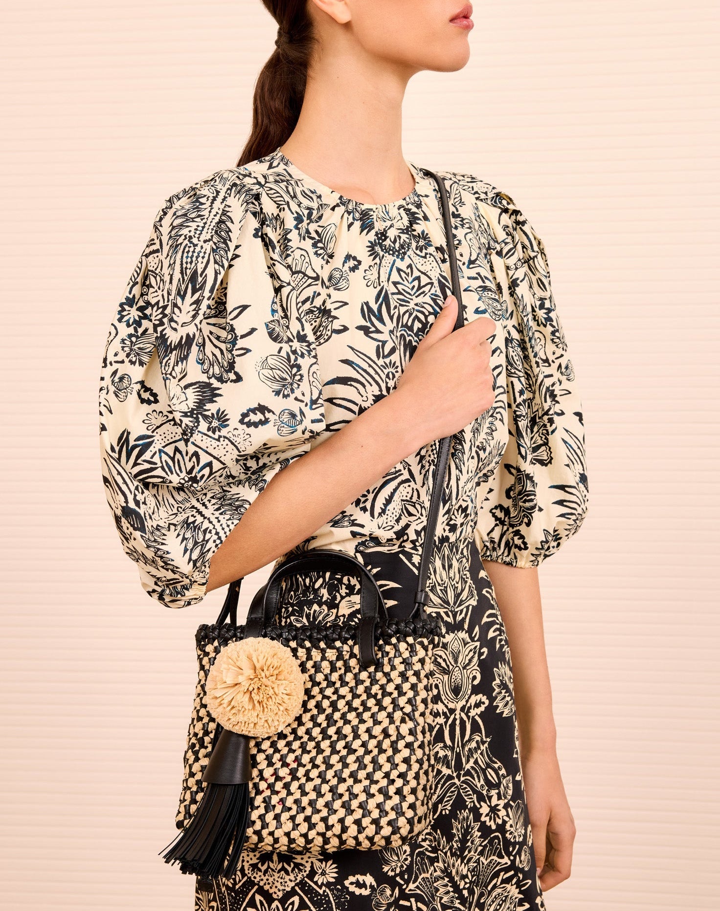 Woman in a floral blouse and skirt holds the Ulla Johnson Raffia Mini Crossbody, featuring a pom-pom and tassel, against a light striped background.