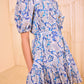 A woman wearing the Ulla Johnson Rosalie Mini Dress, a blue and white floral piece with puffed short sleeves, stands in a neutral-toned room.