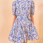 A woman wears the Ulla Johnson Rosalie Mini Dress, a short-sleeved, knee-length style with graphic blue and white botanicals, cinched at the waist with a matching fabric tie.