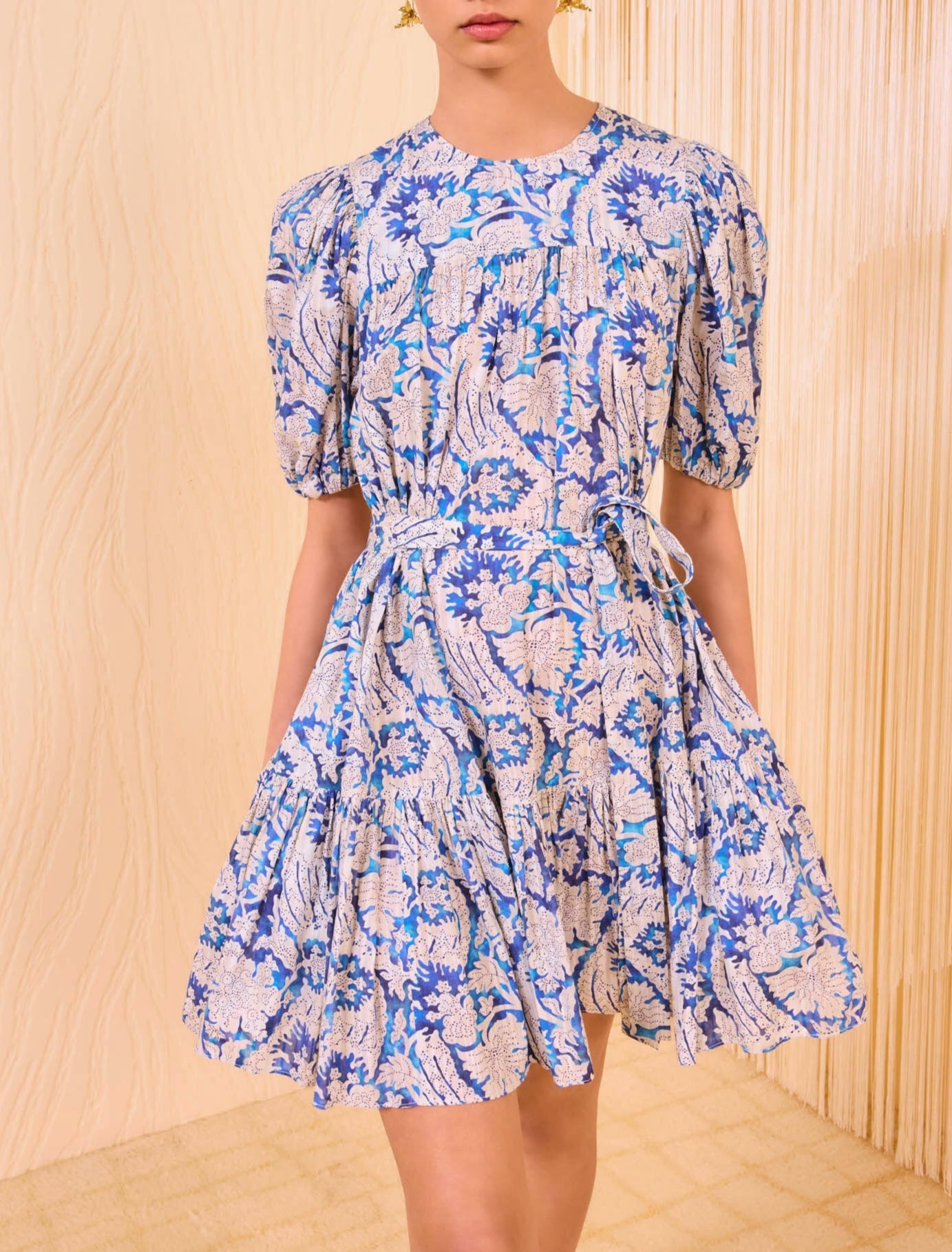 A woman wears the Ulla Johnson Rosalie Mini Dress, a short-sleeved, knee-length style with graphic blue and white botanicals, cinched at the waist with a matching fabric tie.
