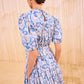 A person with braided hair stands with their back to the camera, wearing the Ulla Johnson Rosalie Mini Dress featuring blue and white graphic botanicals, puff sleeves, and a tiered skirt.