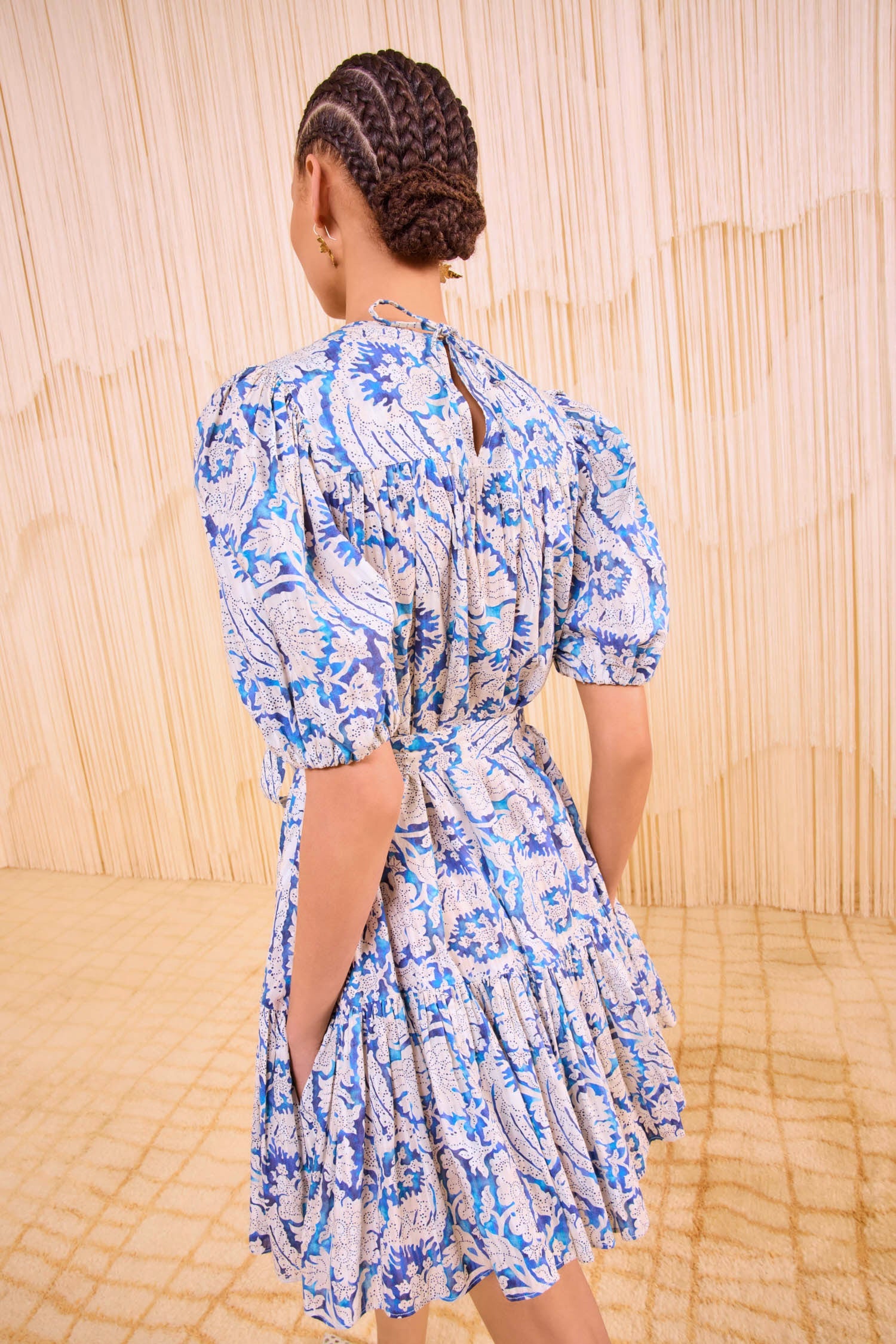 A person with braided hair stands with their back to the camera, wearing the Ulla Johnson Rosalie Mini Dress featuring blue and white graphic botanicals, puff sleeves, and a tiered skirt.