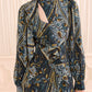 Woman wearing the Sabrina Silk Dress by Ulla Johnson, crafted from luxurious crepe de chine in a blue, gold, and black paisley pattern featuring elegant ruffle details and a diagonal keyhole cutout at the neckline, standing in front of cream vertical paneling.