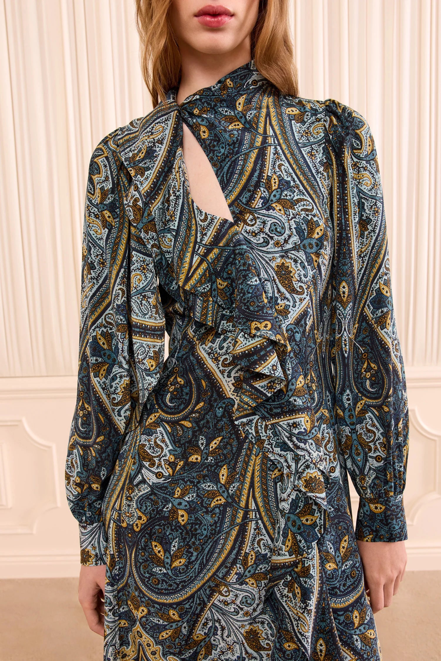 Woman wearing the Sabrina Silk Dress by Ulla Johnson, crafted from luxurious crepe de chine in a blue, gold, and black paisley pattern featuring elegant ruffle details and a diagonal keyhole cutout at the neckline, standing in front of cream vertical paneling.