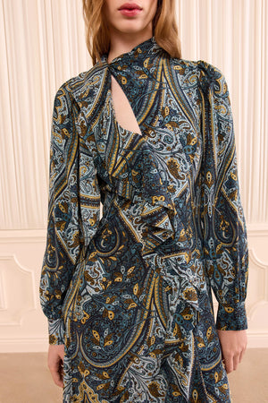 Woman wearing the Sabrina Silk Dress by Ulla Johnson, crafted from luxurious crepe de chine in a blue, gold, and black paisley pattern featuring elegant ruffle details and a diagonal keyhole cutout at the neckline, standing in front of cream vertical paneling.