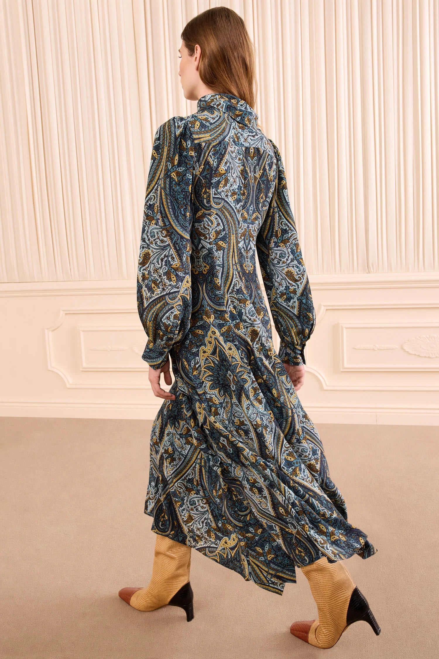 A woman wearing a long-sleeved, ankle-length Sabrina Silk Dress by Ulla Johnson with subtle ruffle details and tan heeled boots walks away in a beige room with vertical wall paneling.