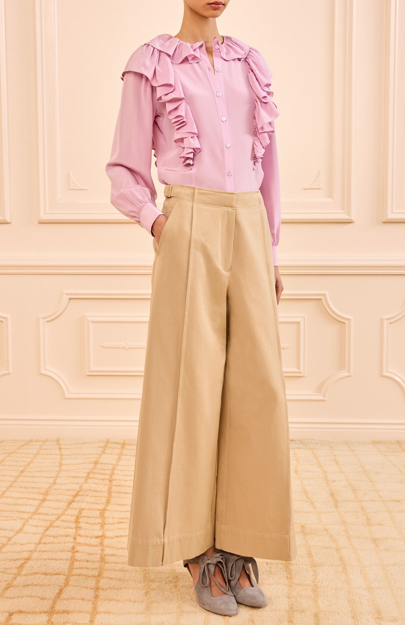A person wears a light pink ruffled blouse tucked into Ulla Johnson's Sascha Cropped Trouser, paired with gray pointed-toe shoes, standing indoors against a cream-colored wall.