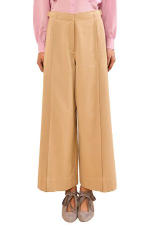 A person stands against a plain background wearing the Ulla Johnson Sascha Cropped Trouser, paired with a light pink long-sleeve shirt and gray ribbon-tied flat shoes.