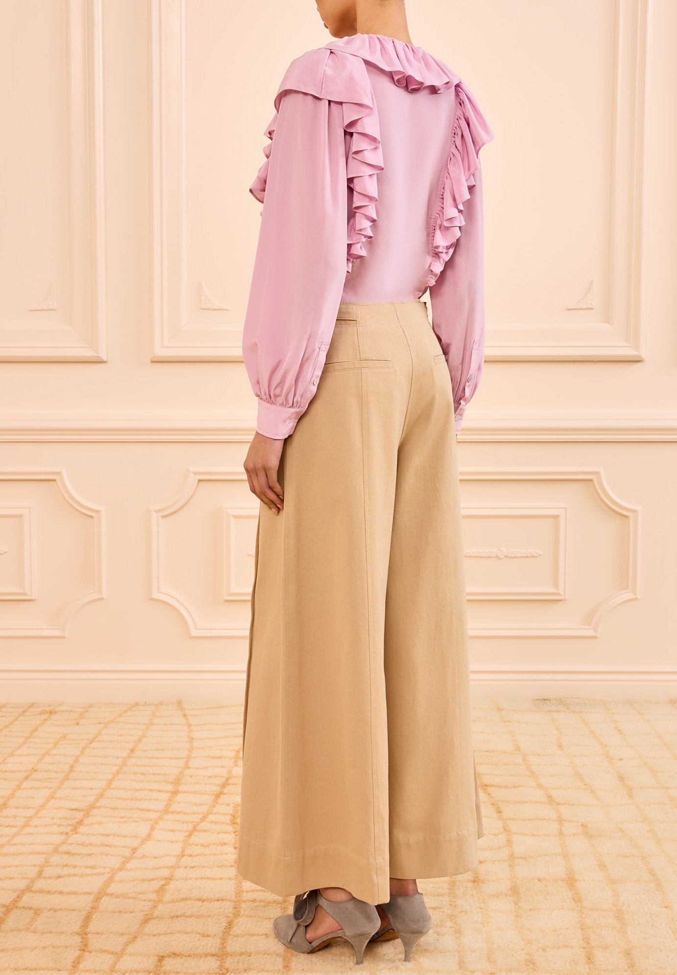 A person stands indoors, seen from behind, wearing a long-sleeved ruffled pink blouse and Ulla Johnson Sascha Cropped Trouser in khaki twill with gray shoes.