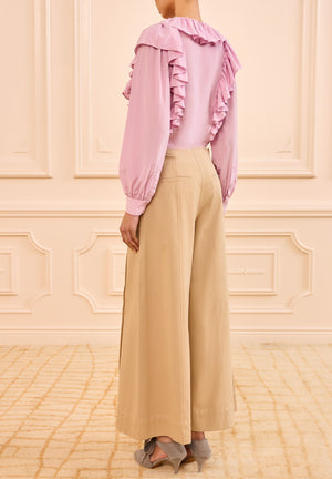A person stands indoors, seen from behind, wearing a long-sleeved ruffled pink blouse and Ulla Johnson Sascha Cropped Trouser in khaki twill with gray shoes.