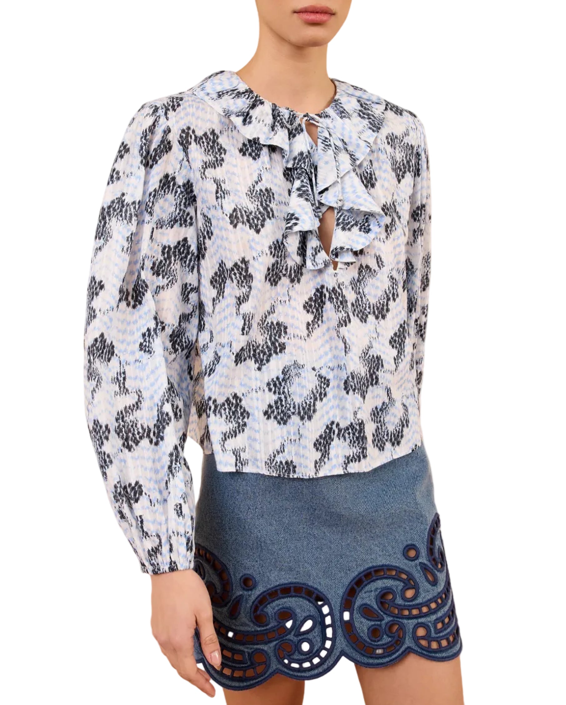 A woman wears the Ulla Johnson Siena Top, a relaxed-fit, long-sleeve patterned blouse with ruffle details, paired with a blue mini skirt featuring decorative cut-out designs at the hem.