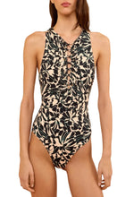 A woman wears the Ulla Johnson Solina One Piece, a black and beige swimsuit featuring vibrant floral motifs and front lace-up detailing, standing against a white background.