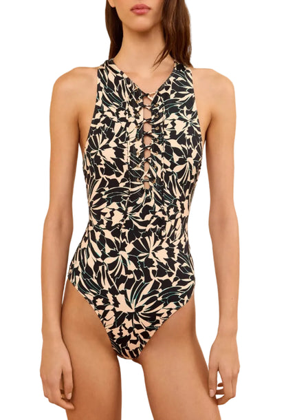 A woman wears the Ulla Johnson Solina One Piece, a black and beige swimsuit featuring vibrant floral motifs and front lace-up detailing, standing against a white background.