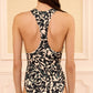 The woman is shown from the back wearing the Ulla Johnson Solina One Piece, a black and beige floral print swimsuit featuring a racerback design and subtle lace-up detailing, while standing indoors against a cream-colored wall.
