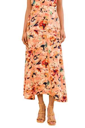 A person is shown from the shoulders down wearing the Ulla Johnson Sophea Skirt—a peach, botanical print cotton poplin skirt with a high-low hemline—paired with tan heeled sandals, set against a white background.