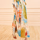 Someone stands indoors on a patterned carpet, dressed in a loose, multicolored Ulla Johnson Studio Dress featuring abstract imagery and short sleeves, paired with a chiffon scarf and brown woven-heeled shoes.