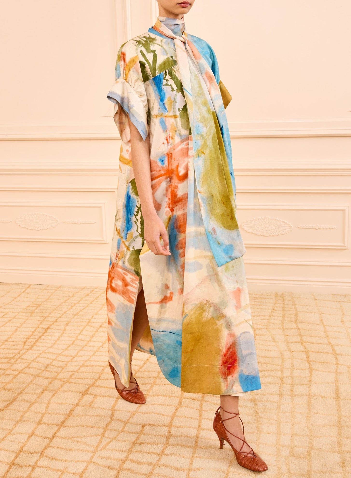 Someone stands indoors on a patterned carpet, dressed in a loose, multicolored Ulla Johnson Studio Dress featuring abstract imagery and short sleeves, paired with a chiffon scarf and brown woven-heeled shoes.