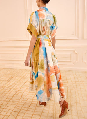 A person faces away from the camera, standing on a beige carpet in a cream room, wearing the Ulla Johnson Studio Dress with abstract patterns, short sleeves, and a belted waist.