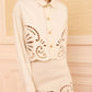 Wearing Ulla Johnson’s The Bobbi Eyelet Jacket—cream-colored and detailed with eyelet embroidery and decorative cut-out patterns along the edges—a person stands against a beige textured wall.