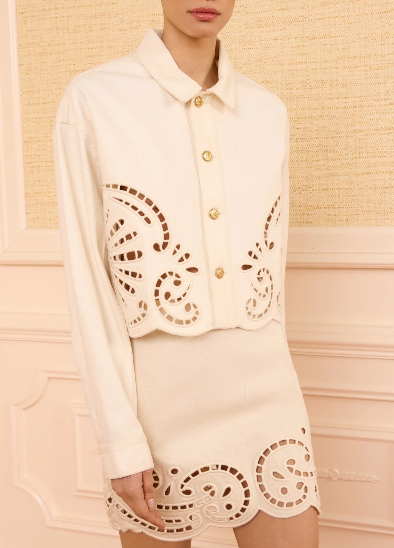 Wearing Ulla Johnson’s The Bobbi Eyelet Jacket—cream-colored and detailed with eyelet embroidery and decorative cut-out patterns along the edges—a person stands against a beige textured wall.