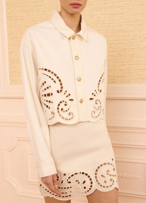 Wearing Ulla Johnson’s The Bobbi Eyelet Jacket—cream-colored and detailed with eyelet embroidery and decorative cut-out patterns along the edges—a person stands against a beige textured wall.