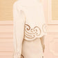 Person facing away, wearing the cream-colored Ulla Johnson Bobbi Eyelet Jacket with delicate laser-cut swirl embroidery along the edges, standing in a pastel-toned paneled room—a modern look that softly merges traditional details with contemporary style.