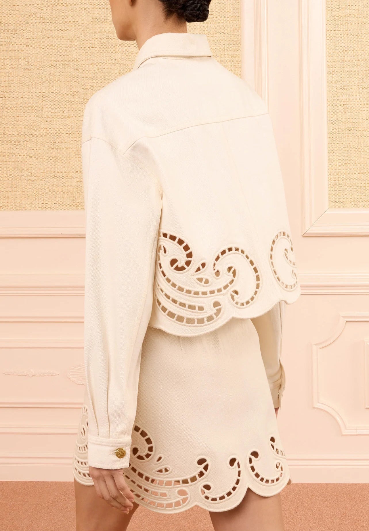 Person facing away, wearing the cream-colored Ulla Johnson Bobbi Eyelet Jacket with delicate laser-cut swirl embroidery along the edges, standing in a pastel-toned paneled room—a modern look that softly merges traditional details with contemporary style.