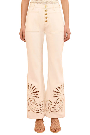 A woman wears the Lou Eyelet Jean by Ulla Johnson—high-waisted white jeans featuring front pockets, gold buttons, and decorative eyelet embroidery near the hems—paired with strappy sandals.