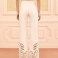 A woman stands with her back to the camera in a beige-furnished room, wearing the Ulla Johnson Lou Eyelet Jean—high-waisted white pants detailed with vintage-inspired eyelet embroidery at the flared hems—and tan heels.