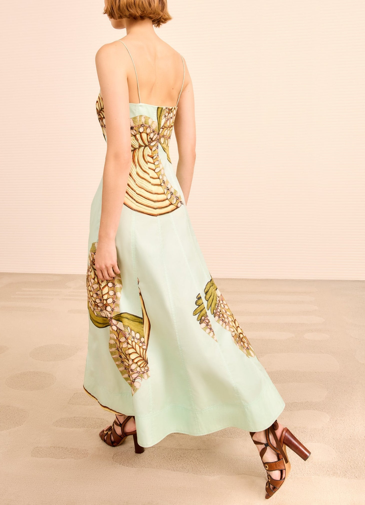 A woman stands on a light floor wearing the Ulla Johnson Vesela Cami Midi Dress—long, light green with a pineapple print and thin straps—paired with brown high-heeled sandals, inspired by Anna Zemánková’s imaginative style.