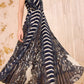 A woman wears the Ulla Johnson Violeta Pleated Midi Dress, featuring intricate dark feather paisley patterns and vertical stripes, as she stands on a patterned carpet before a textured wall.