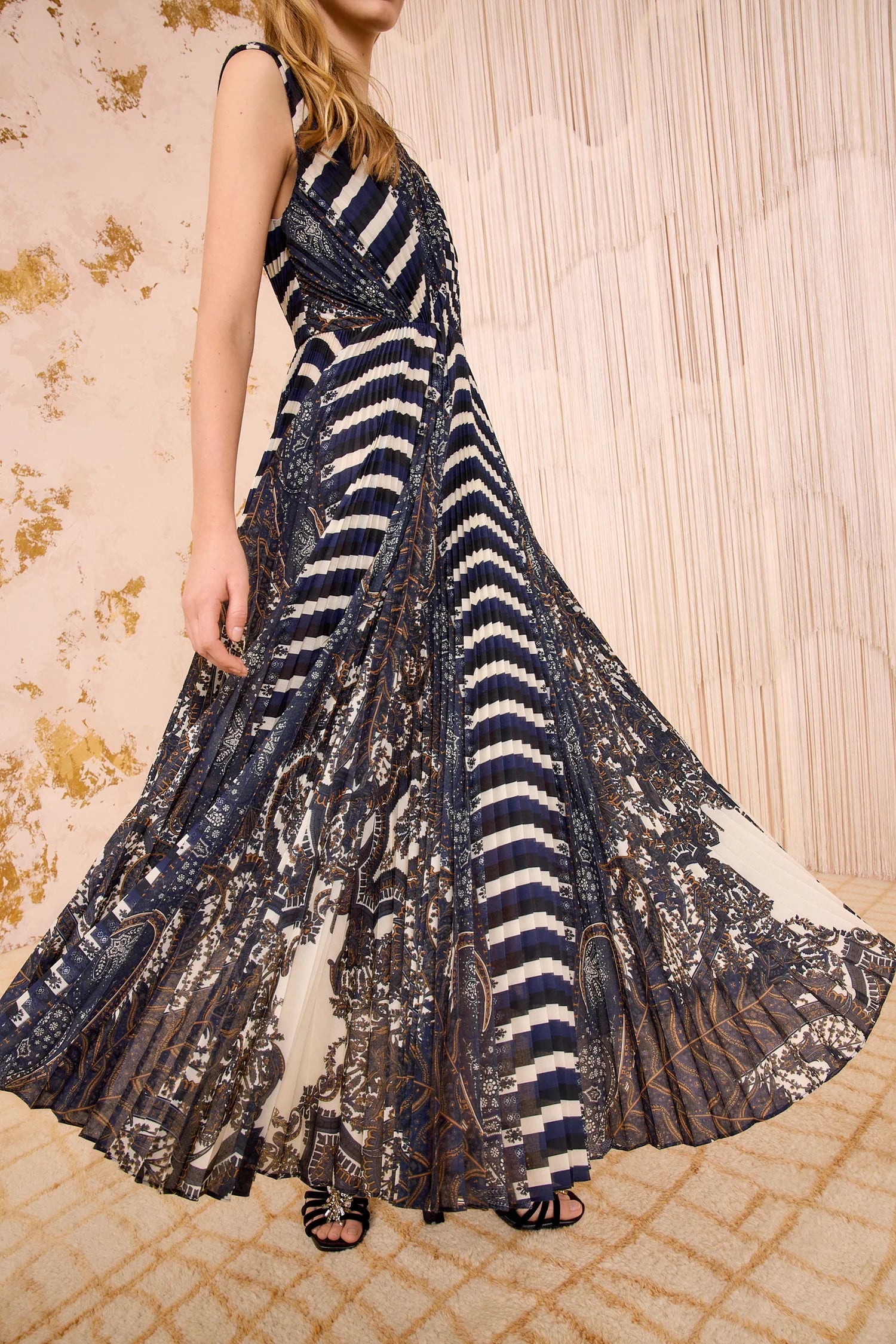 A woman wears the Ulla Johnson Violeta Pleated Midi Dress, featuring intricate dark feather paisley patterns and vertical stripes, as she stands on a patterned carpet before a textured wall.