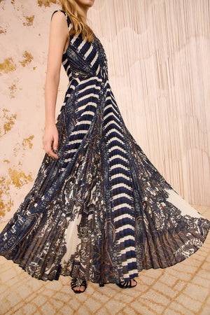A woman wears the Ulla Johnson Violeta Pleated Midi Dress, featuring intricate dark feather paisley patterns and vertical stripes, as she stands on a patterned carpet before a textured wall.