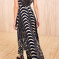 A woman wearing the Violeta Pleated Midi Dress by Ulla Johnson, a sleeveless, multicolored paisley dress with striped patterns, walks on a patterned carpet.