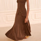 A woman wears the Yanni Strapless Knit Top by Ulla Johnson—a corset-inspired brown knit piece with button details—paired with a matching long skirt and tan boots, standing in a beige room with decorative white paneling.