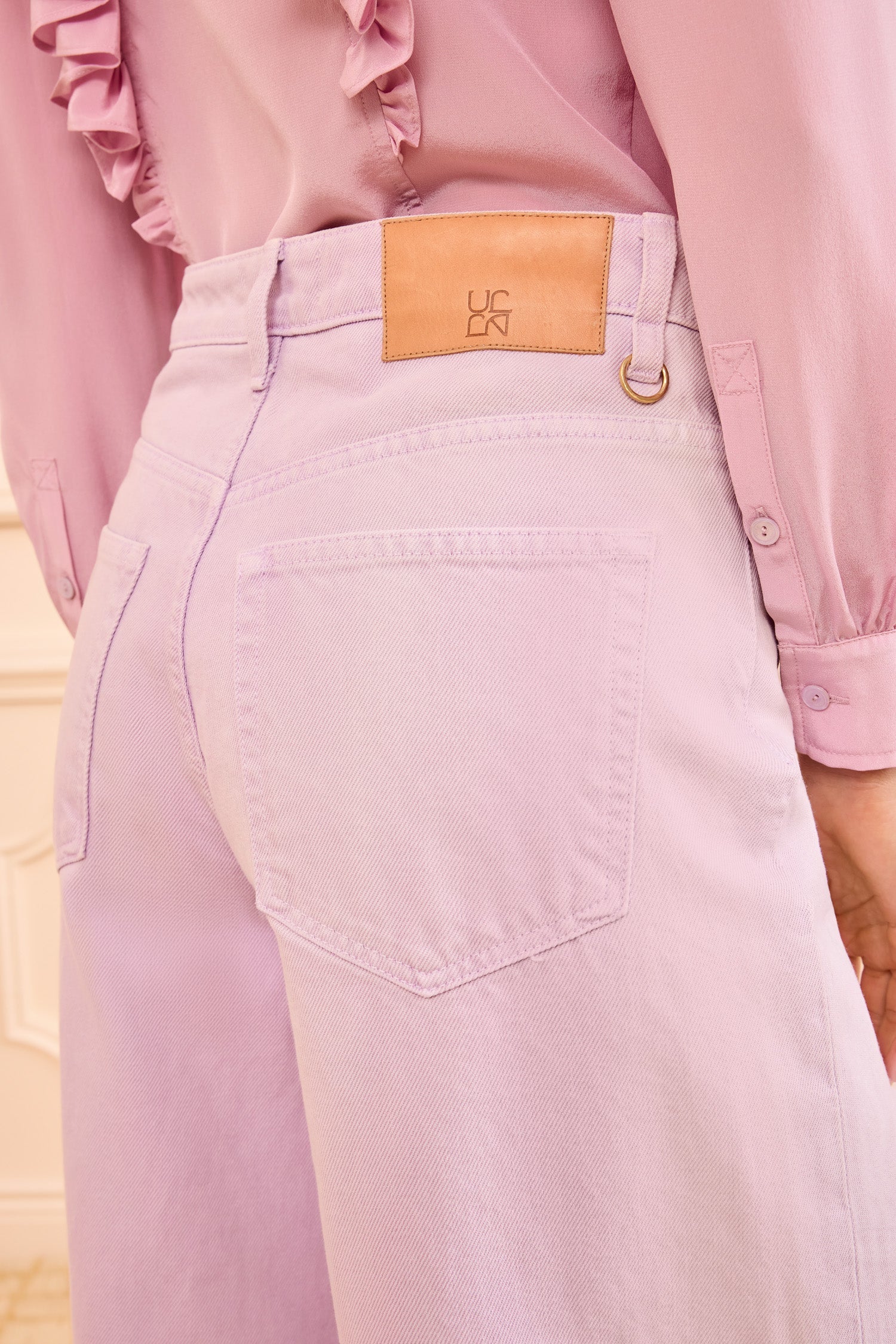 A person wears the Ulla Johnson Zephyr Jean—high-waisted, lavender wash wide-leg jeans with a tan leather patch—paired with a pink ruffle-detail blouse.