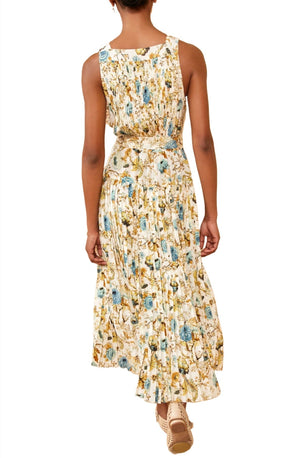 Person wearing an Ulla Johnson Annabeth Dress with 19th century French florals, viewed from the back. The sleeveless dress with pleats and a belt showcases blue, yellow, and brown flowers on silk crepe de chine. Tan-colored sandals are also visible.