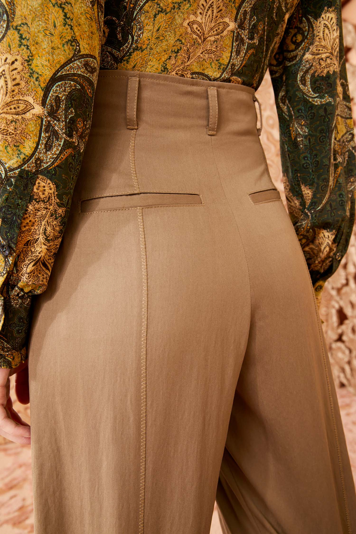 A person wearing Ulla Johnson Delvine Pant in beige, paired with a patterned green and yellow blouse, stands with their back facing the camera.