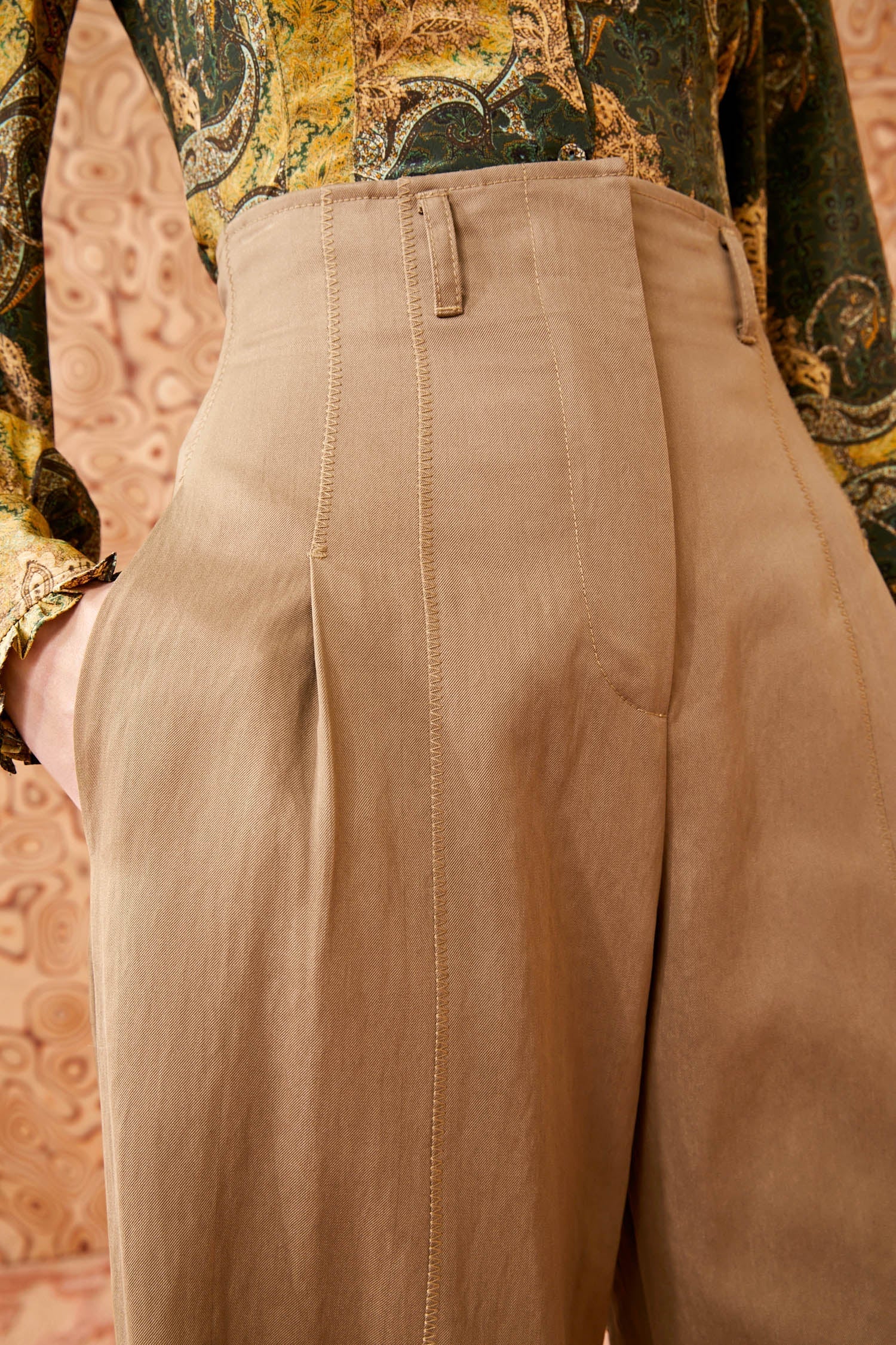 Close-up of a person wearing the stylish Ulla Johnson Delvine Pant in high-waisted beige, paired with a patterned green and yellow shirt. The background features a paisley-like pattern.