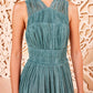 A woman stands before a cream-textured backdrop, wearing Ulla Johnson's sleeveless, pleated teal Freesia Gown featuring a high waist and flowing silhouette.