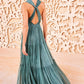 A woman with dark hair in a low ponytail wears the Ulla Johnson Freesia Gown—a teal, floor-length crinkle chiffon dress with a crossed back and cutout—standing before an ornate beige backdrop.