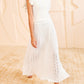 A person wears the Ulla Johnson Giulia Top, featuring pleats and eyelet details, paired with white sandals, standing indoors on a patterned floor.