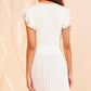 A woman with curly hair is seen from behind wearing the Ulla Johnson Giulia Top, an elegant white blouse with pleated details and ruffled sleeves, set against a peach-colored textured wall.